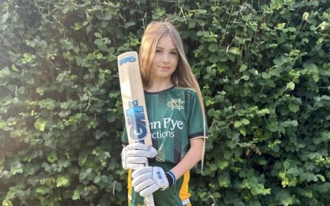 A girl with long blonde hair, wearing a green cricket uniform and holding a cricket bat